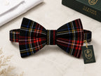 A pre-tied bow tie with a black, red, and blue tartan plaid pattern, displayed on a light marble surface next to its green Glenor Heritage packaging box.