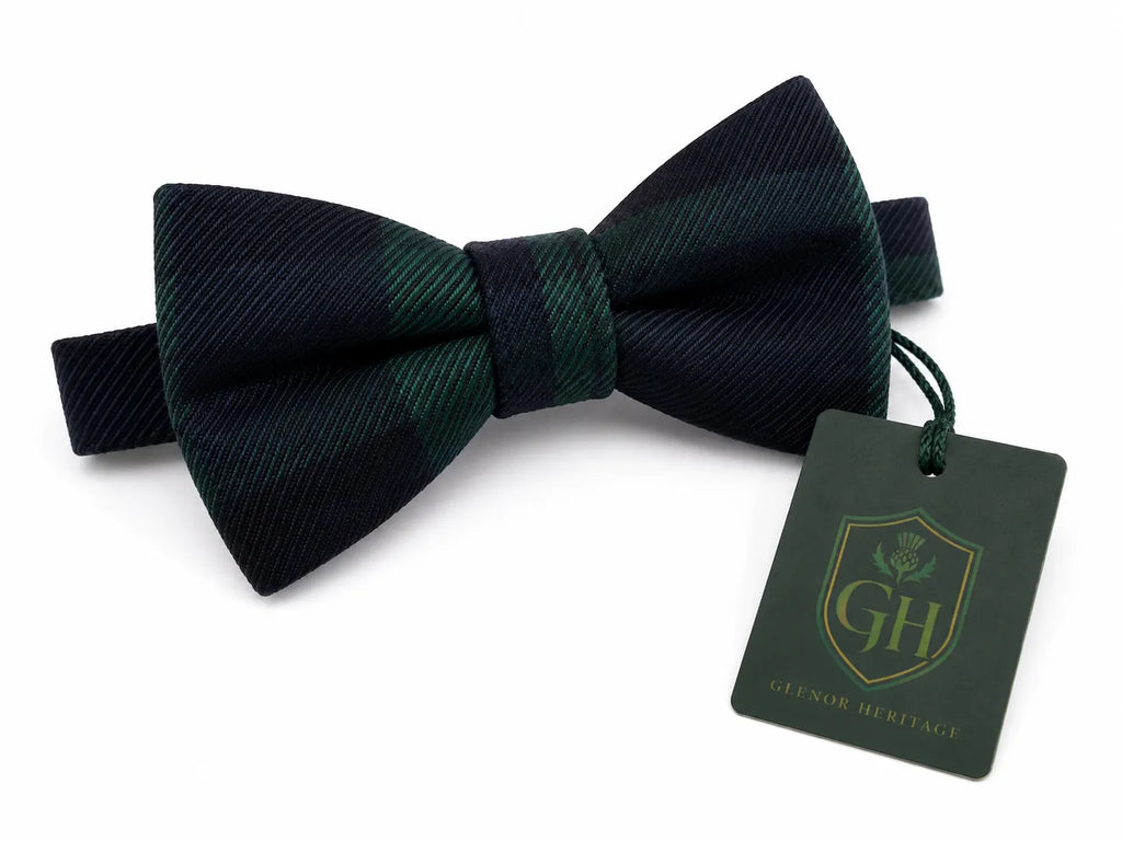 A dark green and black tartan plaid bow tie with a matching green hangtag featuring a gold crest logo for 'Glenor Heritage'.
