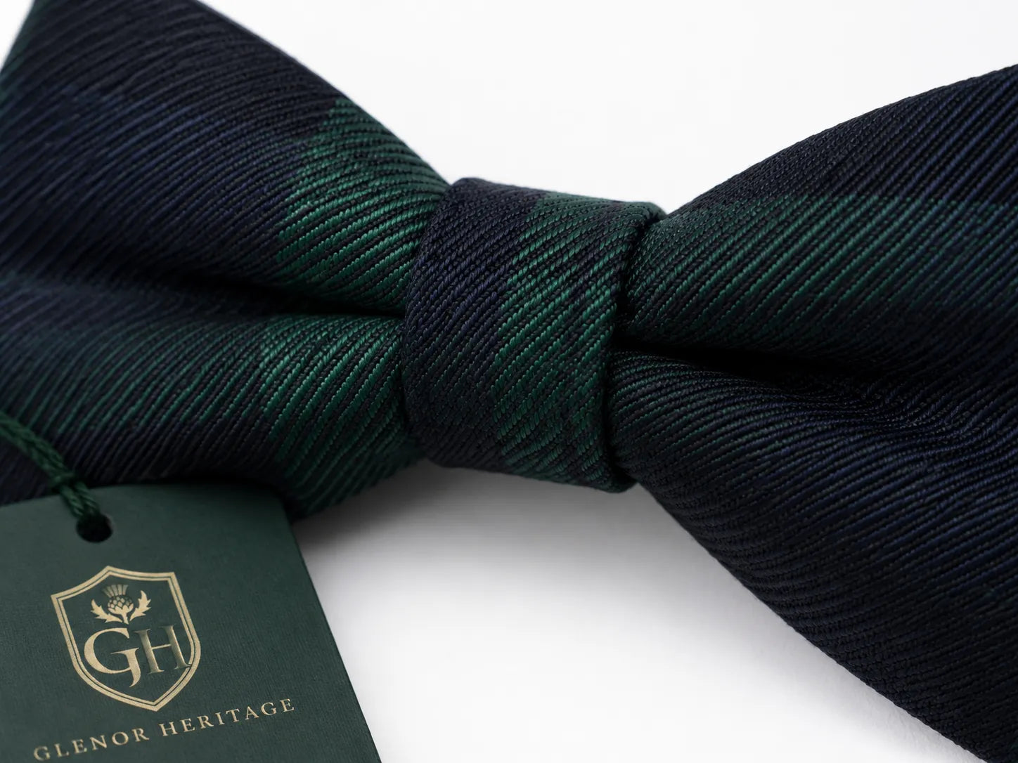 A dark green and navy plaid bow tie with a matching dark green paper brand tag featuring the Glenor Heritage logo, set against a clean white background.