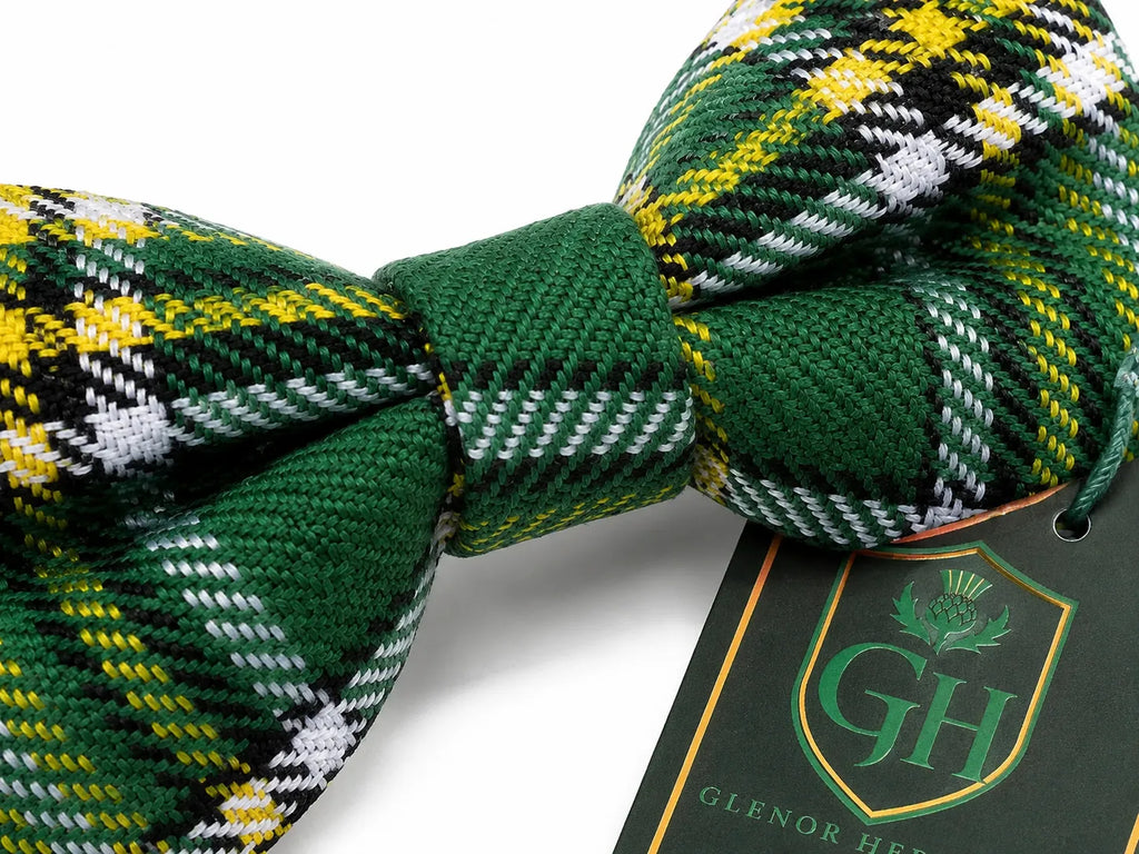 A green tartan plaid bow tie with yellow, white, and black stripes, featuring a dark green paper tag with a 'GH Glenor Heritage's logo.