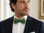 A man wearing a dark navy suit jacket, white dress shirt, and a green tartan plaid bow tie, shown in a close-up portrait.