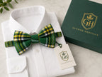 A green and yellow tartan plaid bow tie styled on a white dress shirt collar, next to a dark green Glenor Heritage gift box and branded paper tag.