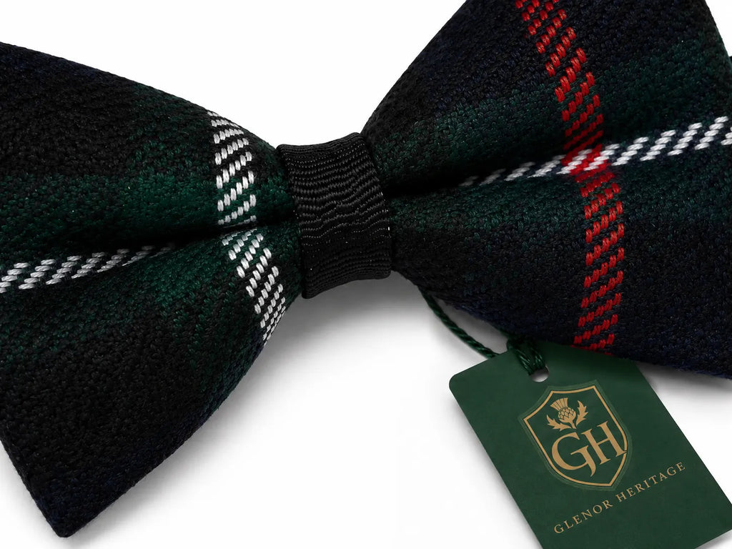 A dark green and black plaid bow tie with red and white stripe details, tied with a black ribbon and featuring a green 'Glenor Heritage' brand tag.