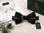 A pre-tied bow tie in a dark green and black tartan plaid pattern with red and white stripes, displayed on a marble surface next to a white dress shirt, a dark green Glenor Heritage box, and a small thistle sprig. A green hangtag with the Glenor Heritage logo is attached to the bow tie.