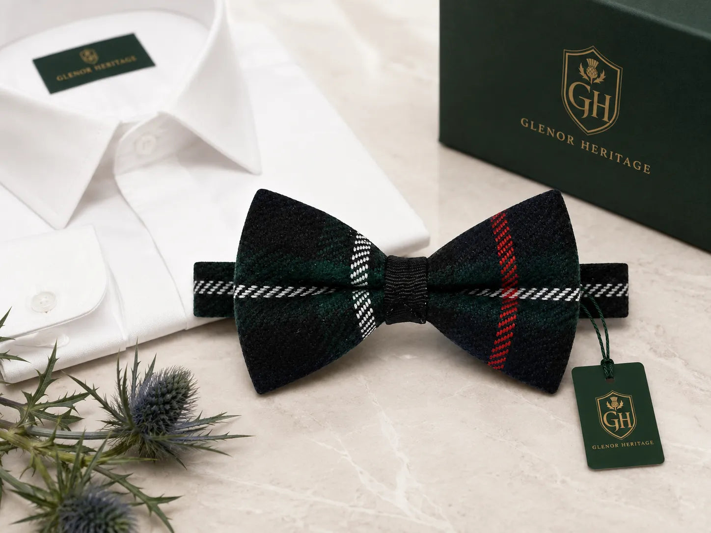 A pre-tied bow tie in a dark green and black tartan plaid pattern with red and white stripes, displayed on a marble surface next to a white dress shirt, a dark green Glenor Heritage box, and a small thistle sprig. A green hangtag with the Glenor Heritage logo is attached to the bow tie.