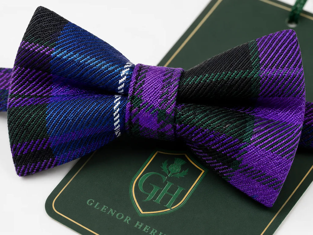A pre-tied bow tie with a purple, black, and green tartan plaid pattern, displayed on a dark green branded card that reads 'GLENOR HERITAGE'.