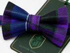 A pre-tied bow tie with a purple, black, and green tartan plaid pattern, displayed on a dark green branded card that reads 'GLENOR HERITAGE'.