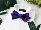 A white dress shirt folded neatly with a purple and black plaid bow tie tied at the collar. A green Glenor Heritage gift box, ribbon, and branded hangtag are arranged around it on a light marble surface with greenery in the corner.